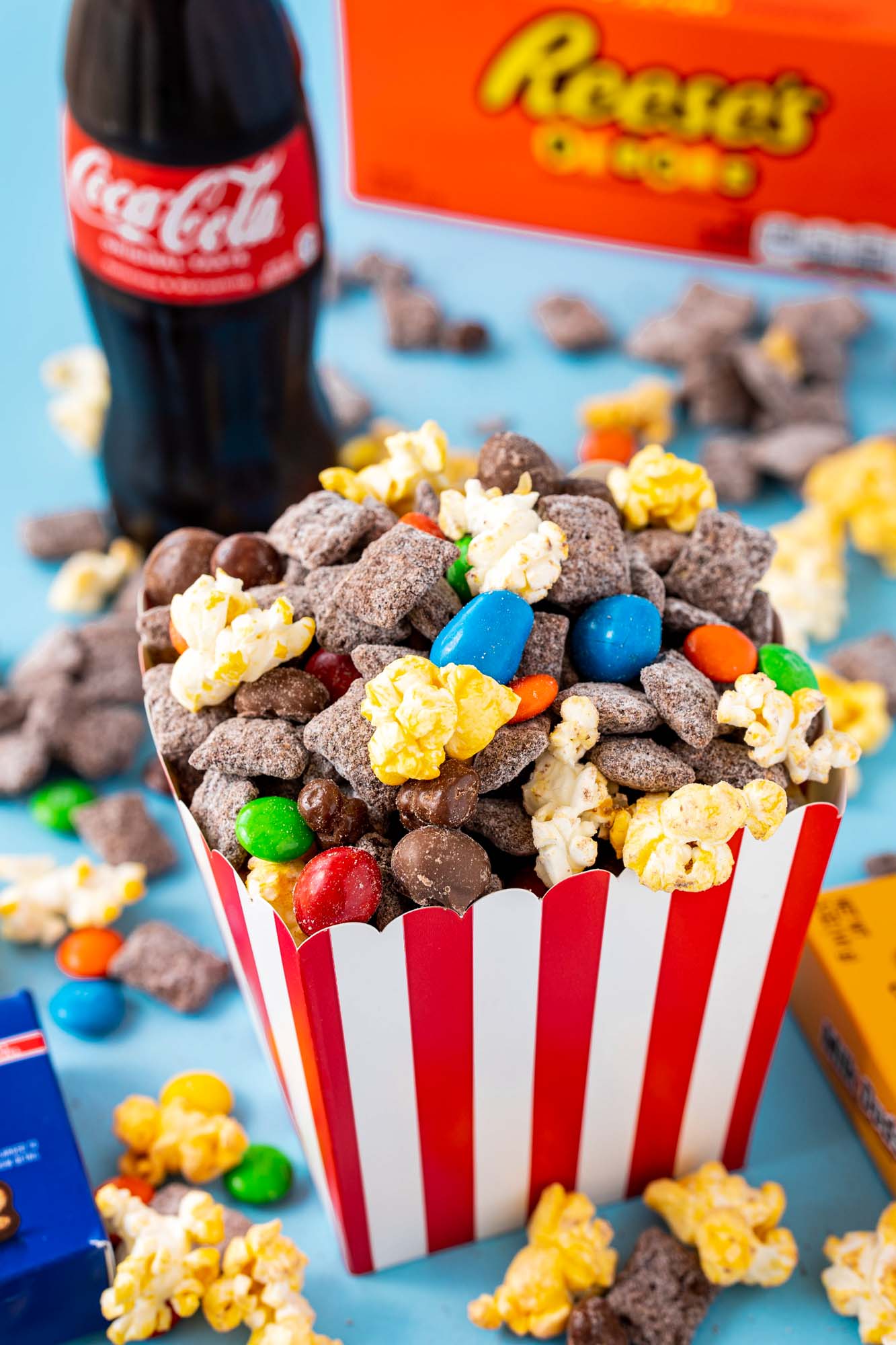 a small red and white striped popcorn box filled with move theatre muddy buddy snack mix. A coca cola bottle is in the background along with a box of reeses pieces