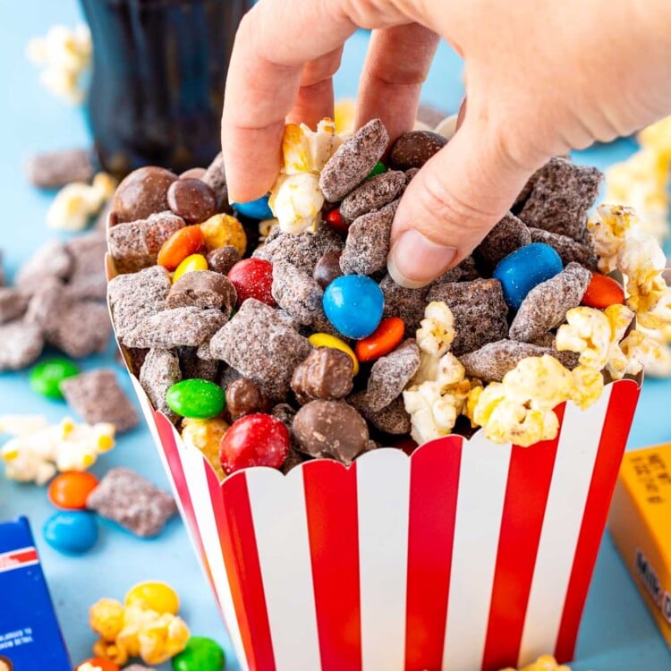 a human hand reaching into a red and white popcorn box filled with chocolate chex mix with popcorn and candy.