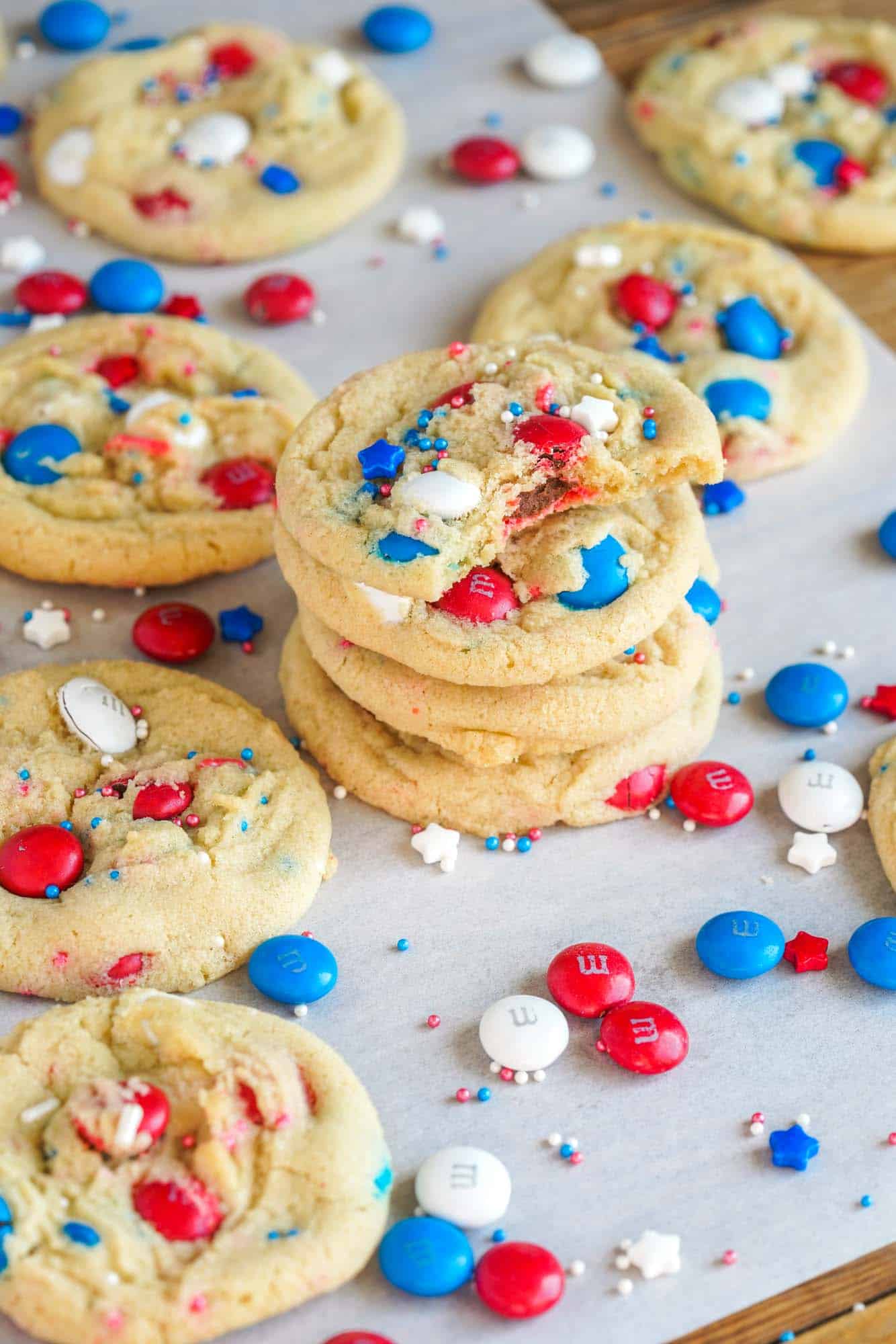 Red White And Blue M&M Cookies on white parchment paper, showing a bite shot. With patriotic M&M's and sprinkles scattered.