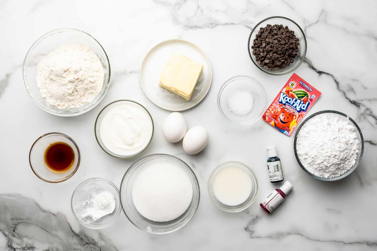 The ingredients needed to make watermelon cupcakes with kool aid buttercream, all measured into small bowls, on a marble counter and viewed from above.