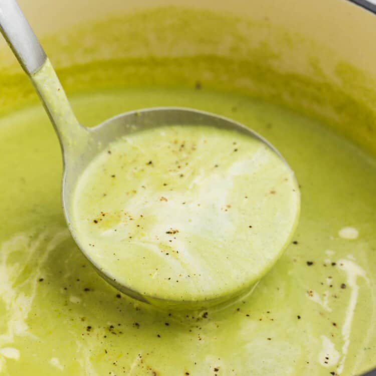 a ladle serving creamy, green zucchini soup from a red dutch oven.