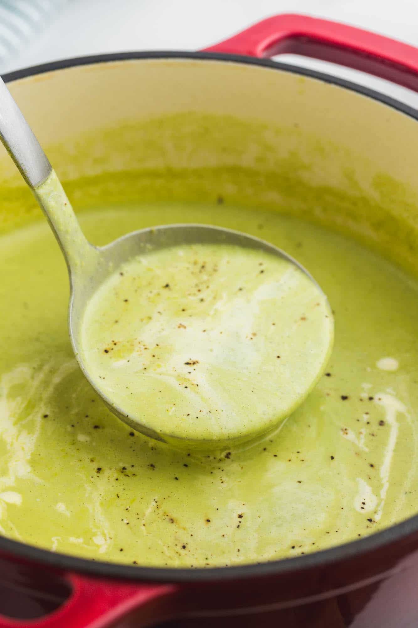 a ladle serving creamy, green zucchini soup from a red dutch oven.