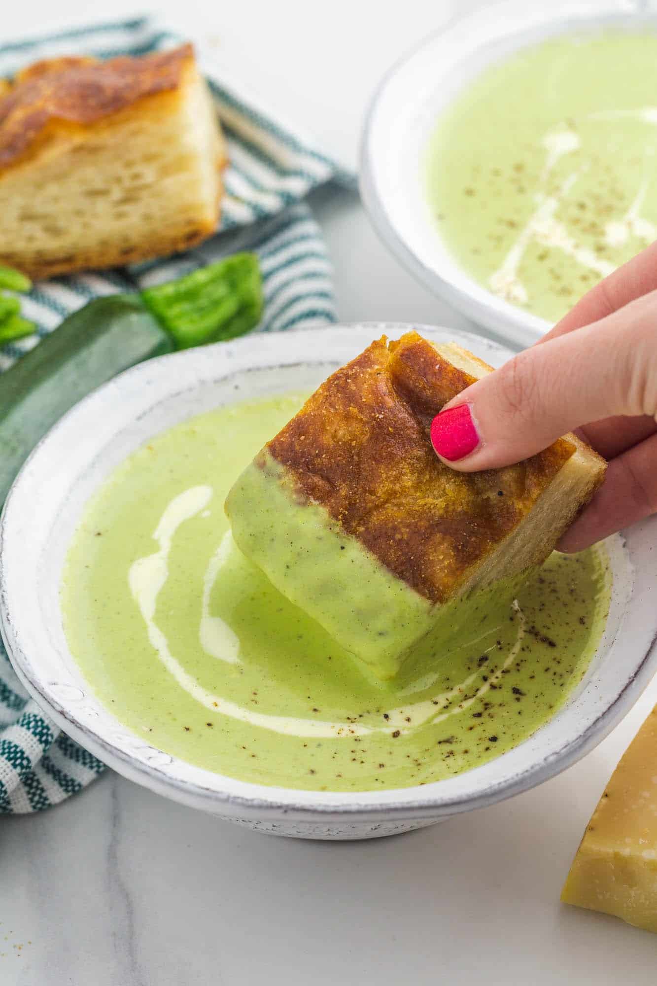 a feminine hand dipping a piece of foccacia into a bowl of creamy zucchini soup