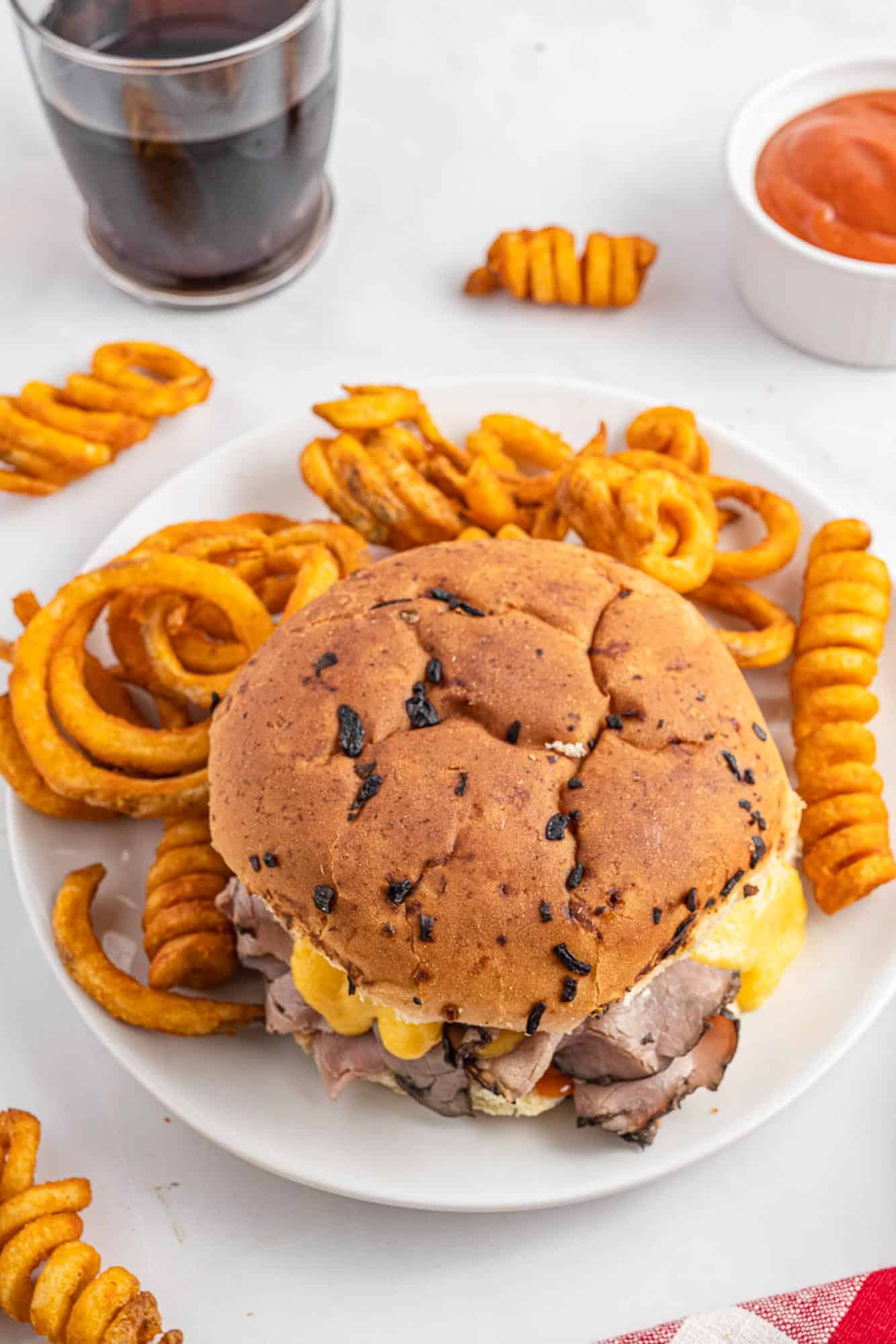 a roast beef and cheddar sandwich on an onion roll on a plate with curly fries