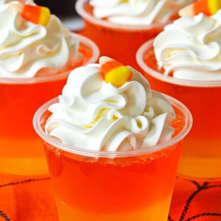 4 orange and yellow layered jello cups topped with whipped cream and candy corn. Text at bottom of image says Candy Corn Jello Cups