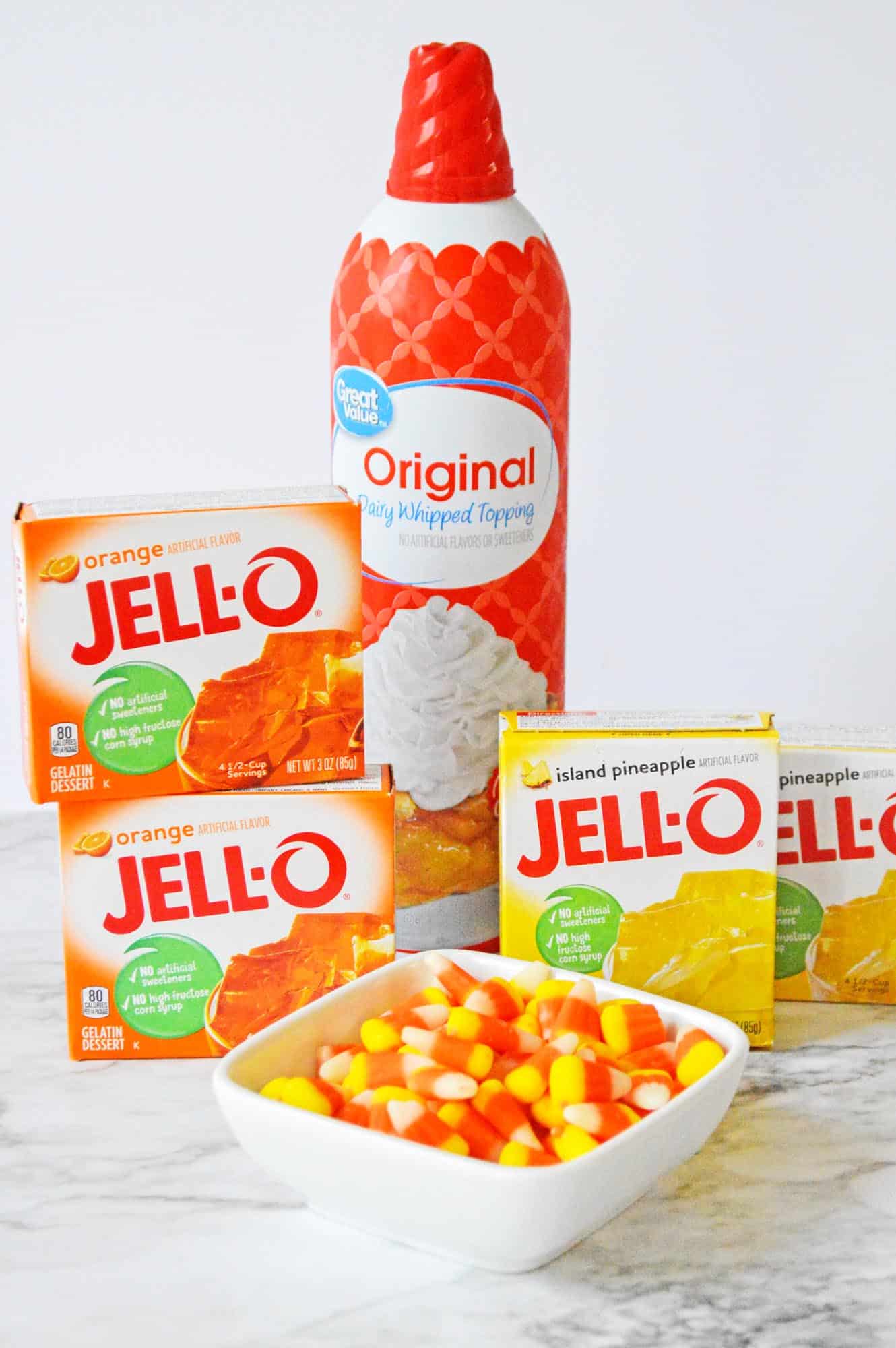boxes of orange and pineapple jello, a can of whipped cream, and a bowl of candy corn. 