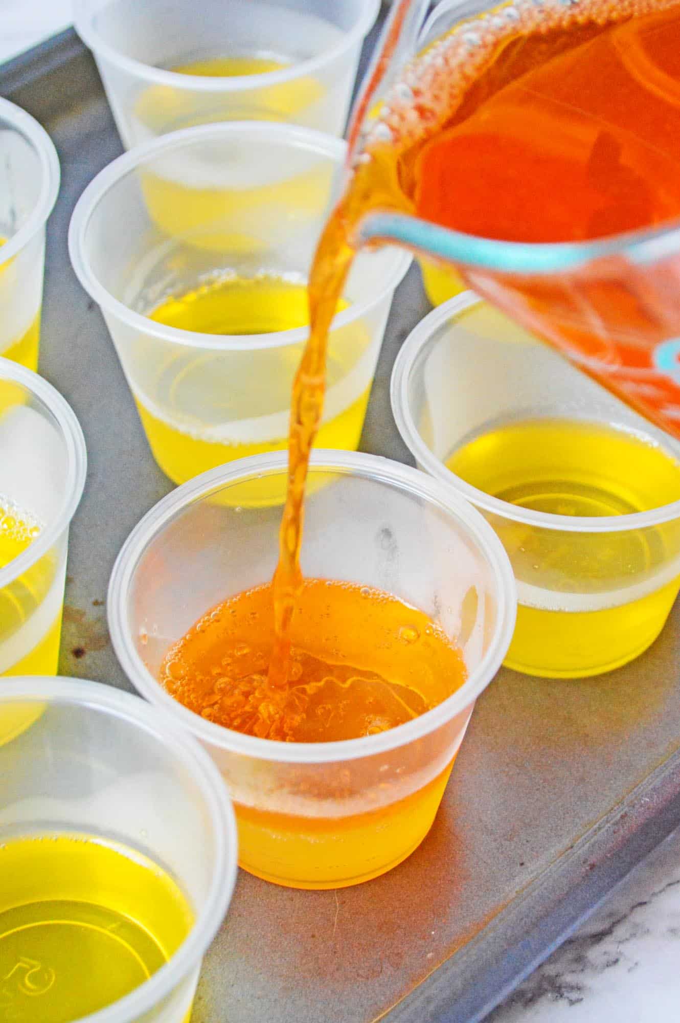 cups half filled with yellow jello. Orange jello is being poured into the cups