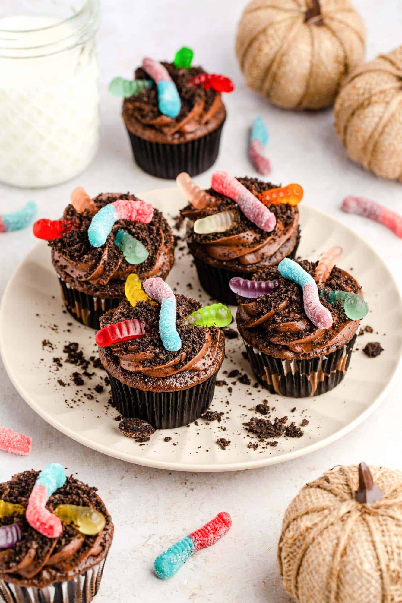 a plate holding four cupcakes with another cupcake on the table behind it. The cupcakes are chocolate with chocolate frosting, topped with oreo crumbs and candy gummy worms in multiple colors