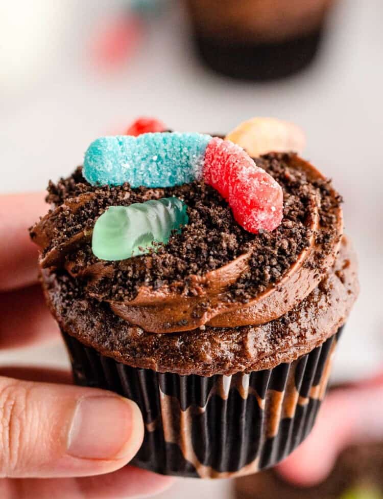 a hand holding a chocolate cupcake topped with chocolate frosting, dirt crumbs, and sour gummy worms