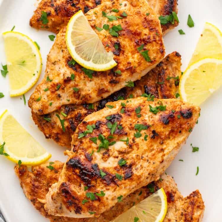 Grilled yogurt marinated chicken cutlets served on a white platter with lemon slices and chopped parsley