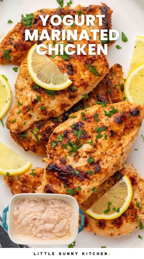 Grilled yogurt marinated chicken cutlets served on a white platter with lemon slices and chopped parsley, and overlay text that says "yogurt marinated chicken"