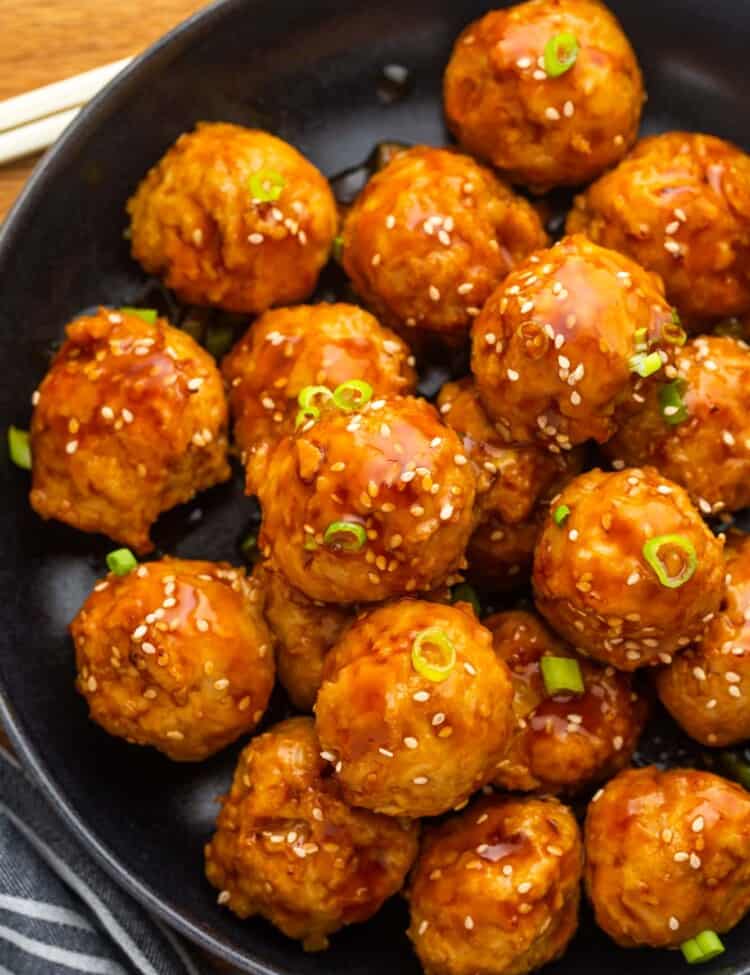 Overhead shot of Asian chicken meatballs in a black plate, garnished with chopped scallions and sesame seeds.