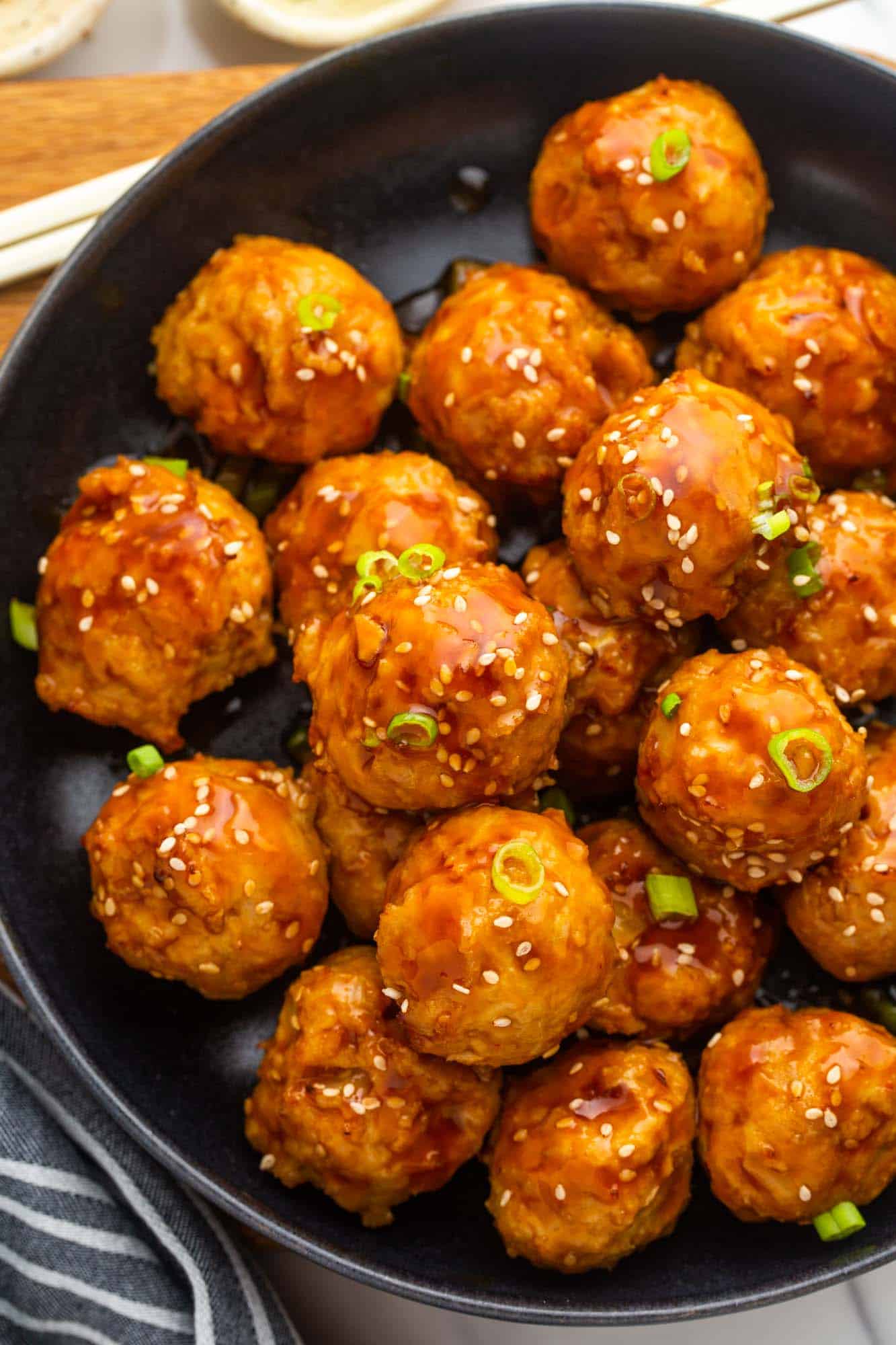 Overhead shot of Asian chicken meatballs in a black plate, garnished with chopped scallions and sesame seeds.