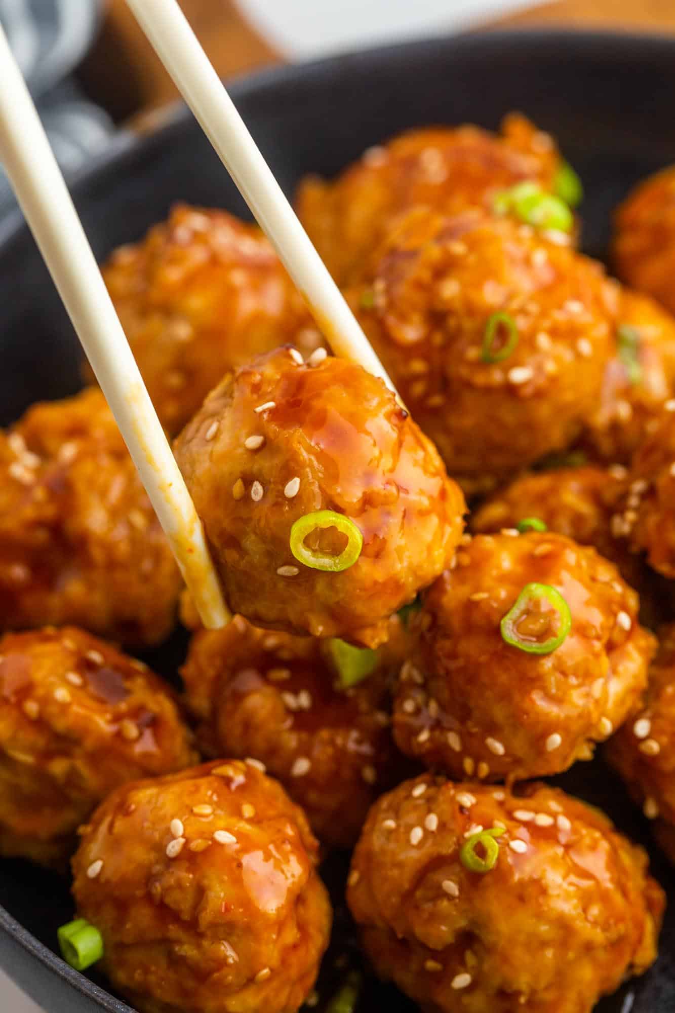 a black bowl of meatballs with asian sauce and sesame seeds. One is being held by chopsticks.