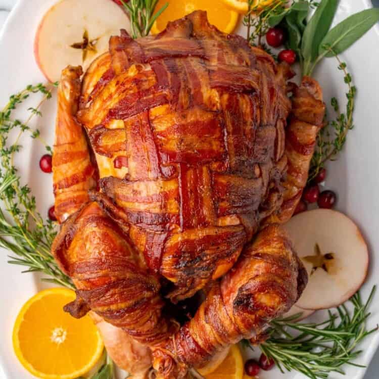 a turkey, completely wrapped in slices of bacon in a cross cross lattice pattern. The turkey is on a platter lined with fresh herbs, cranberries, apple slices, and orange slices.