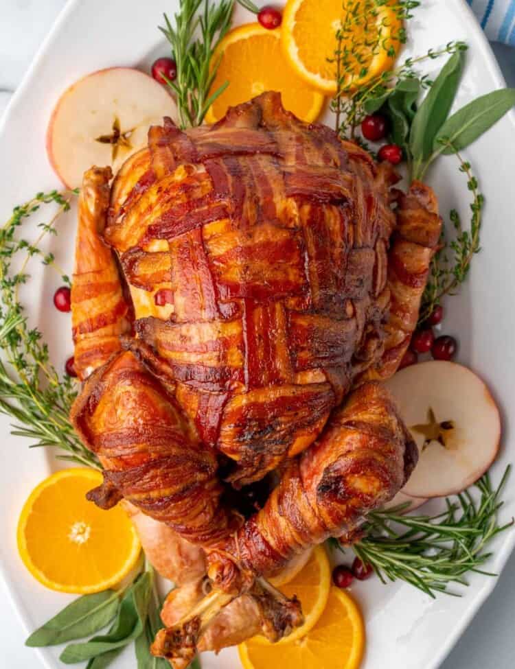a turkey, completely wrapped in slices of bacon in a cross cross lattice pattern. The turkey is on a platter lined with fresh herbs, cranberries, apple slices, and orange slices.