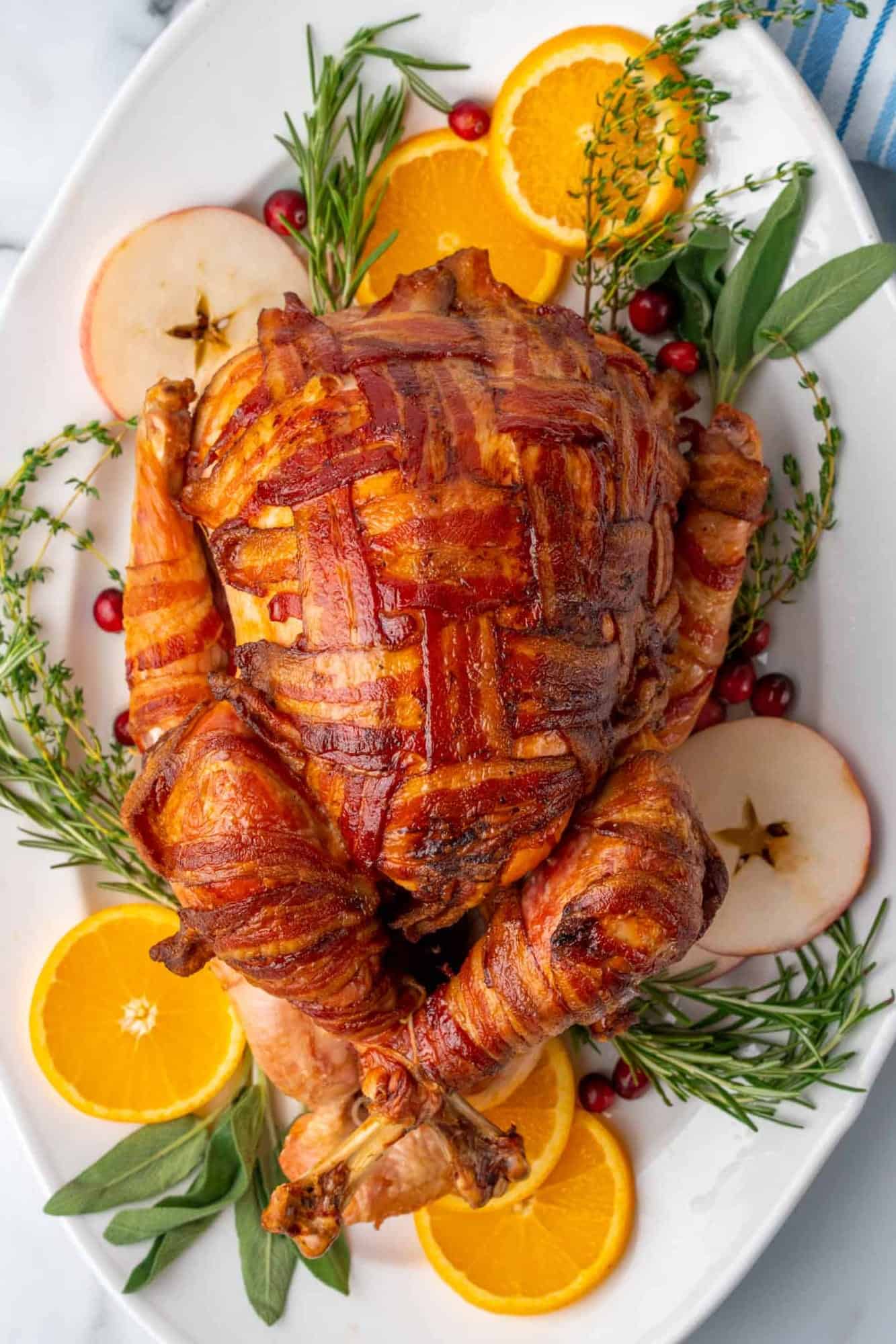 a turkey, completely wrapped in slices of bacon in a cross cross lattice pattern. The turkey is on a platter lined with fresh herbs, cranberries, apple slices, and orange slices.