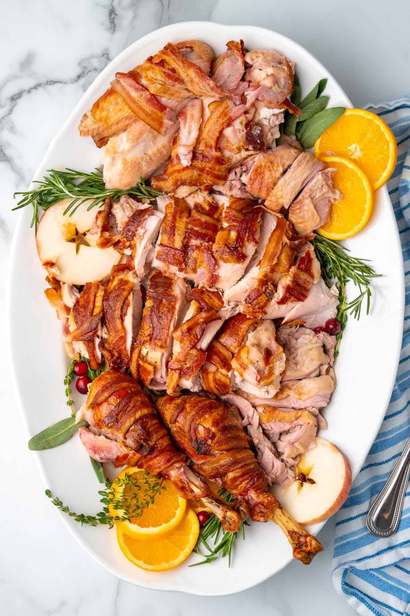 a large platter lined with herbs and fruit slices. A bacon wrapped turkey that has been carved sits on top.