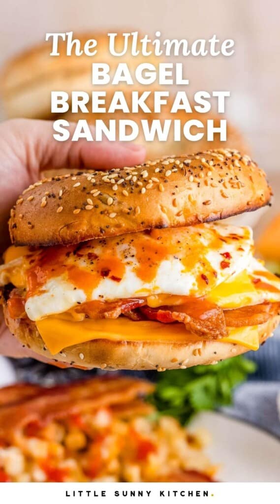 Holding a bagel breakfast sandwich with hot sauce. And overlay text that says "The Ultimate Bagel Breakfast Sandwich"