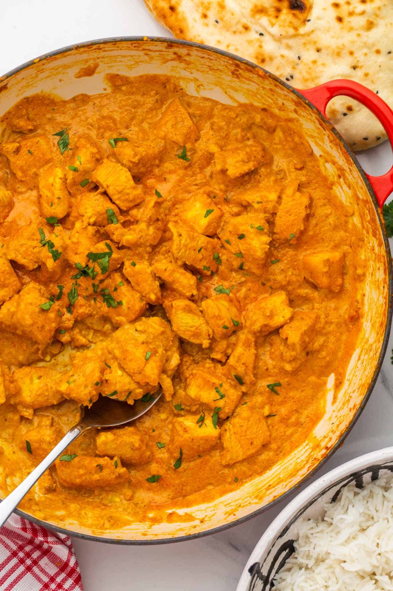 a large red, white enameled skillet filled with chicken korma with a brownish sauce. A spoon is serving from the pan. 