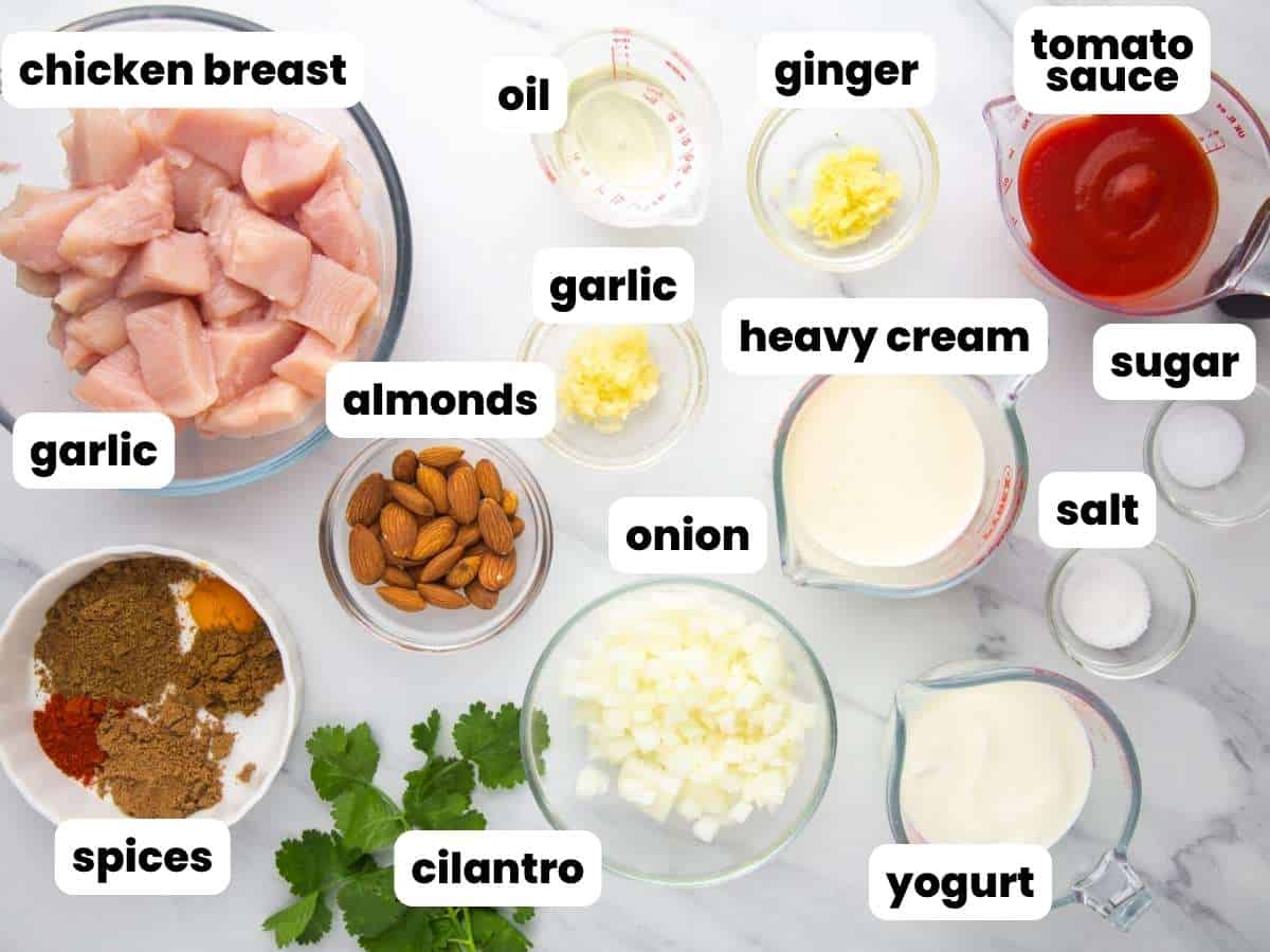 The ingredients needed to make chicken korma at home, all measured into small bowls and arranged on a counter, viewed from above. Small white text boxes label each ingredient. 
