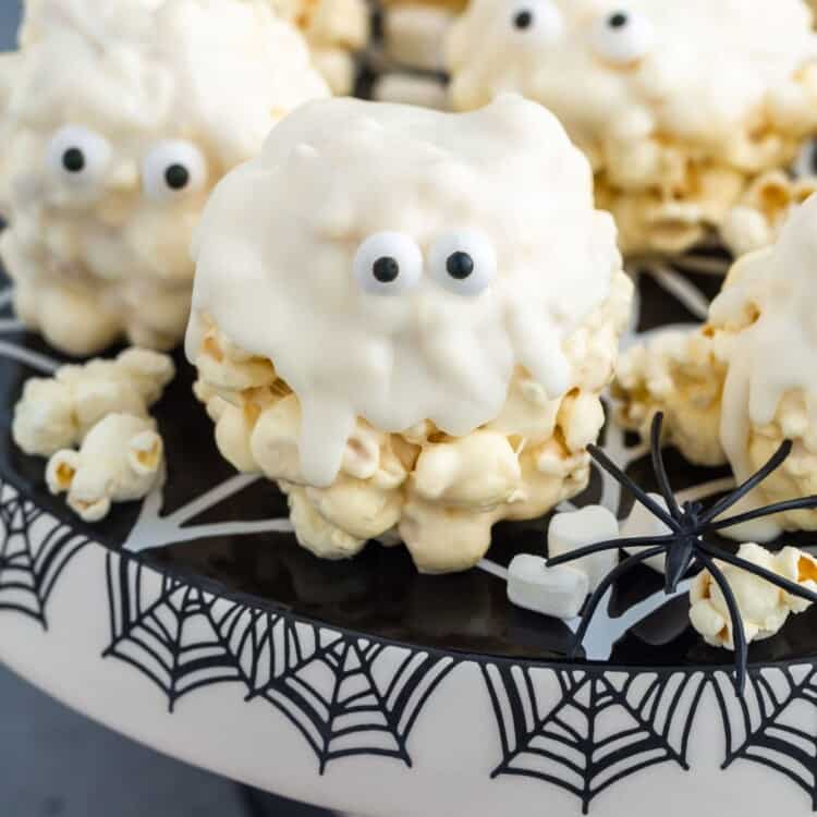 Popcorn balls that are dipped in white chocolate and given candy eyes so that they look cute ghosts. They are on a black and white cake stand decorated with spider webs.