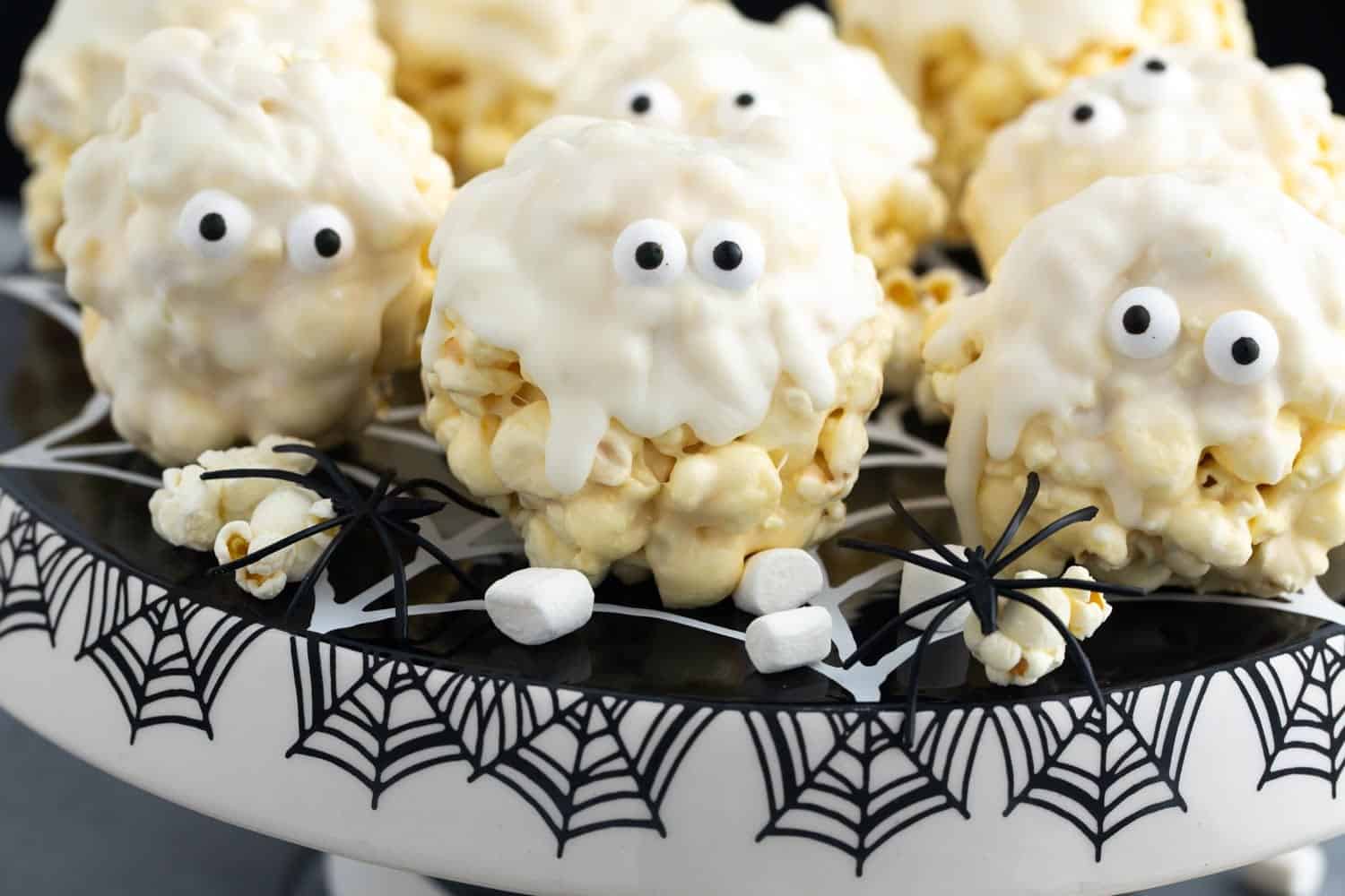 White marshmallow popcorn balls shaped like ghosts with candy eyes, placed on a halloween themed cake stand.