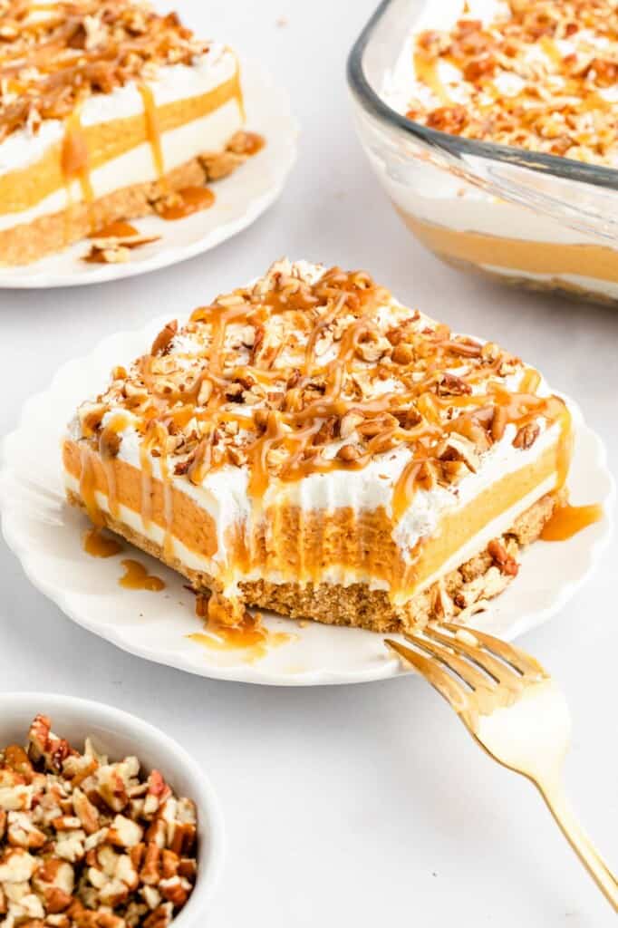 a large square piece of pumpkin lush, a layered lasagna style dessert topped with crushed pecans and caramel. The slice is on a white plate, and a fork is reaching toward it. A bite has been taken from one corner.