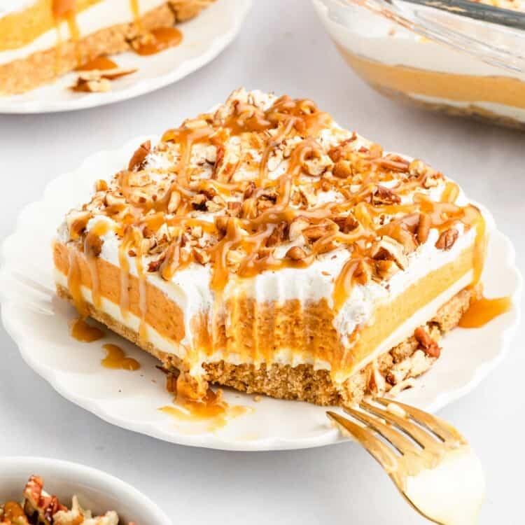 a large square piece of pumpkin lush, a layered lasagna style dessert topped with crushed pecans and caramel. The slice is on a white plate, and a fork is reaching toward it. A bite has been taken from one corner.
