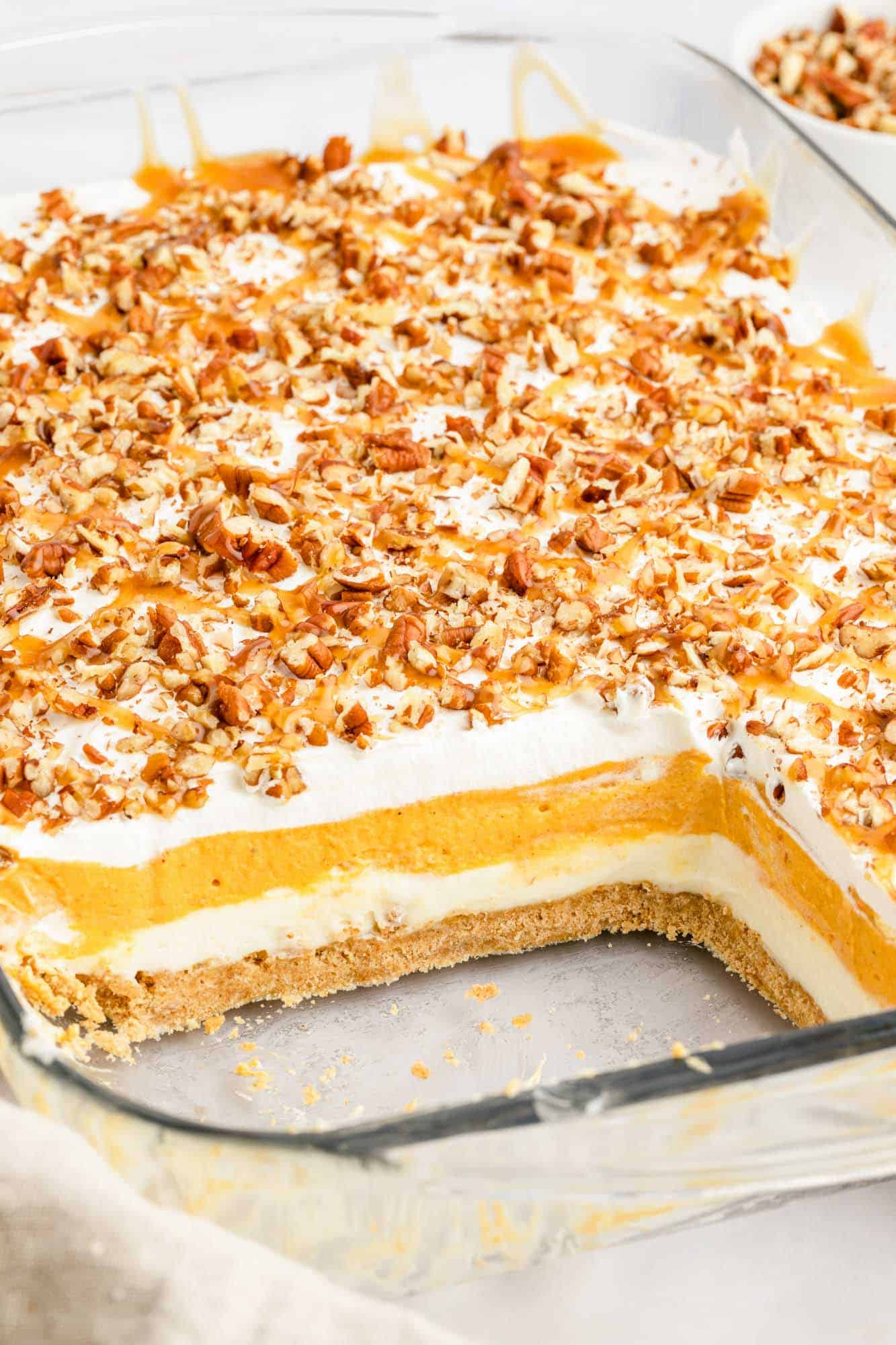 a glass casserole dish filled with no-bake pumpkin lush with layers of white cream, pumpkin cream, a graham cracker crust, and pecan topping. Two square of the dessert have been removed.