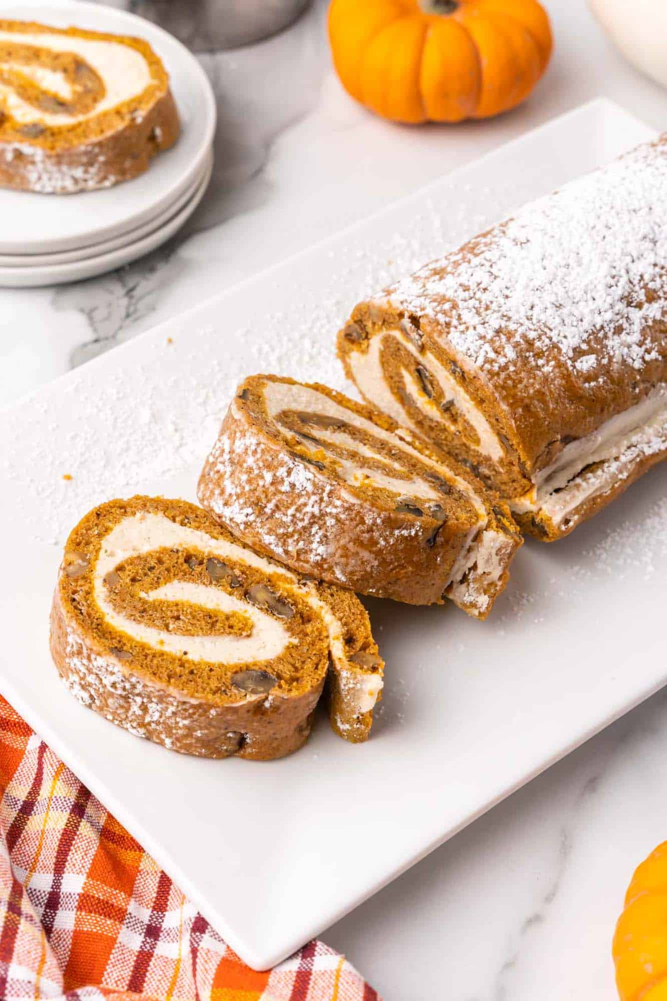 a rectangular white platter holding a pumpkin roll cake sprinkled with powdered sugar. Two thick slices have been made at one end. 