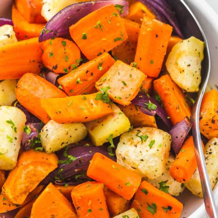 Roasted root vegetables served in a white bowl, with a serving spoon on the side.