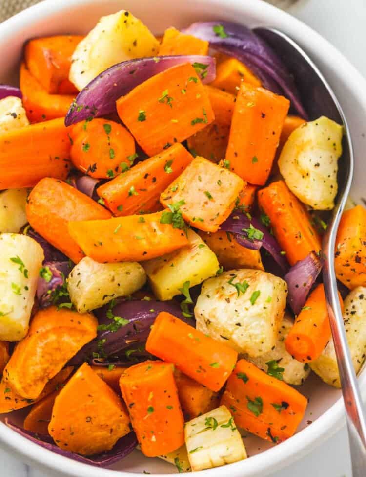 Roasted root vegetables served in a white bowl, with a serving spoon on the side.