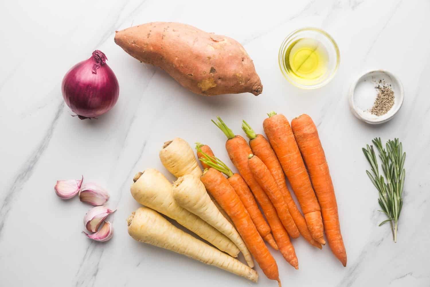 Ingredients needed to make roasted root vegetables including a sweet potato, red onion, carrots, parsnips, garlic, olive oil, rosemary, salt and pepper.