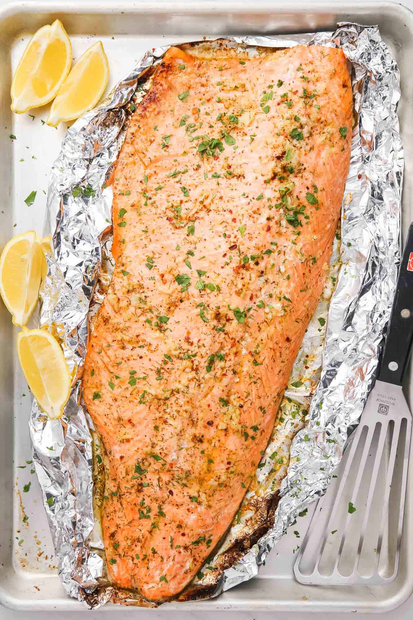 Overhead shot of baked trout fillet on foil, with lemon wedges on the side and a fish spatula
