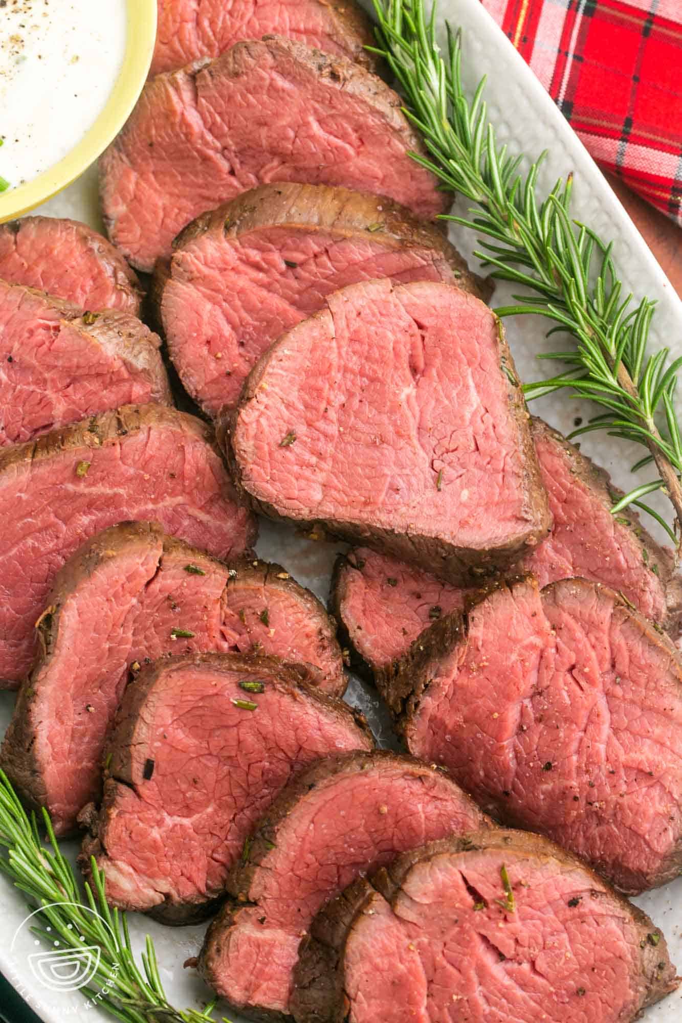 Sliced perfectly rosy beef tenderloin on an oval platter with rosemary sprigs on the sides.
