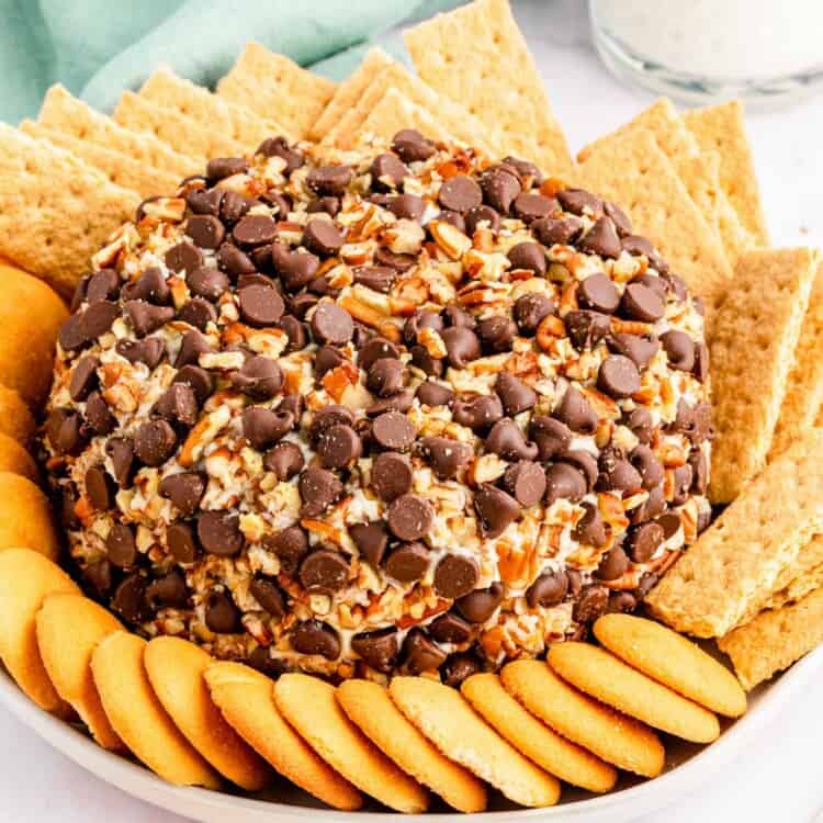 a cream cheese ball with pecans and chocolate chips, surrounded by graham crackers and nilla wafers. a glass of milk and a mint green napkin are in the background.