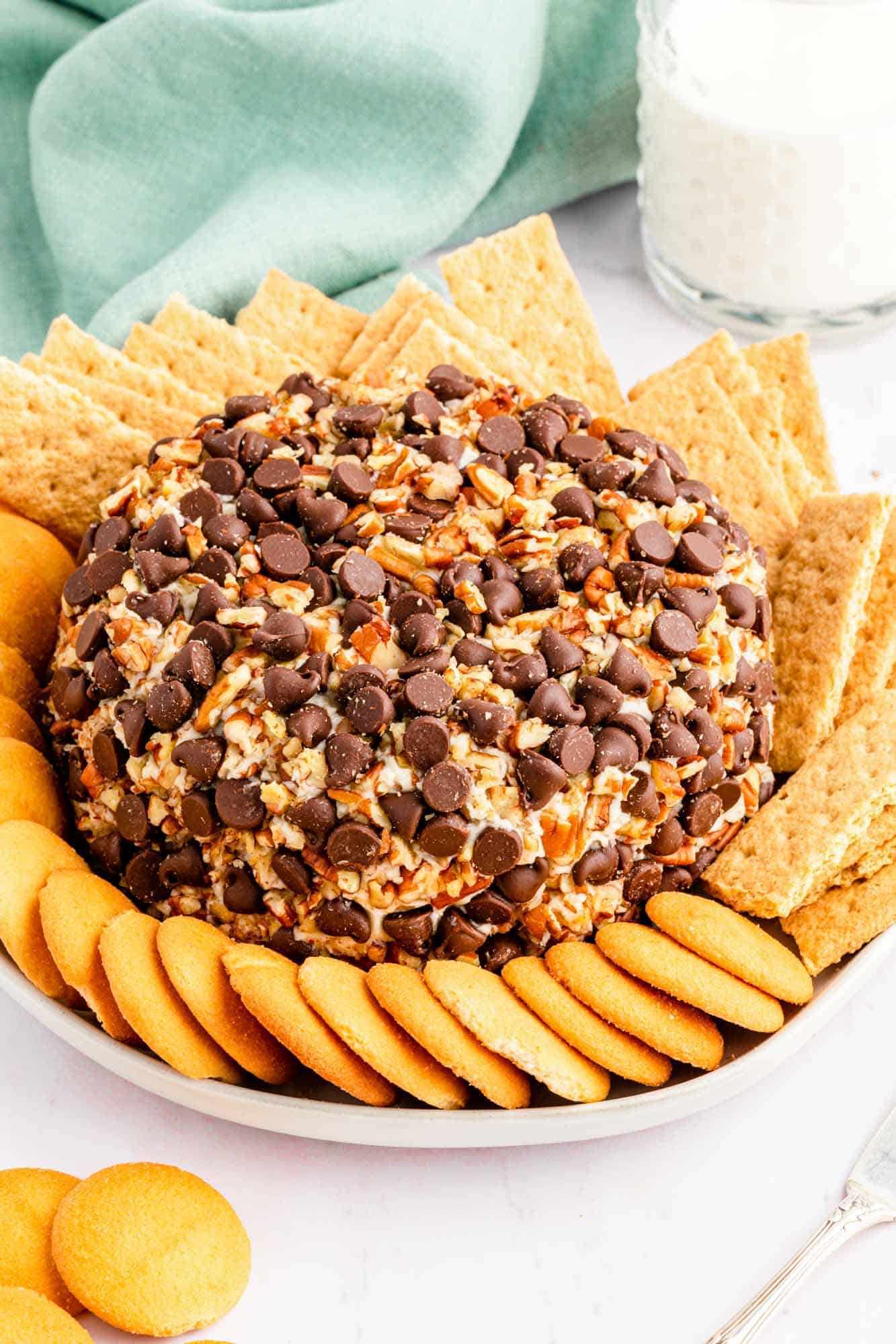 a cream cheese ball with pecans and chocolate chips, surrounded by graham crackers and nilla wafers. a glass of milk and a mint green napkin are in the background. 