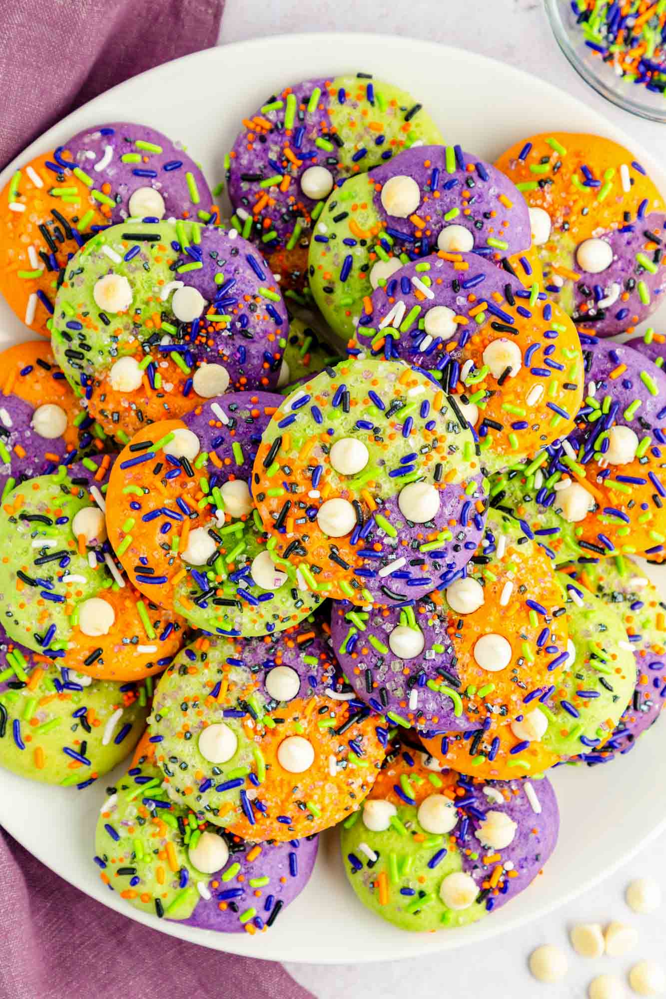a white plate filled with colorful halloween cookies swirled with orange, green, and purple, topped with sprinkles and white chocolate chips
