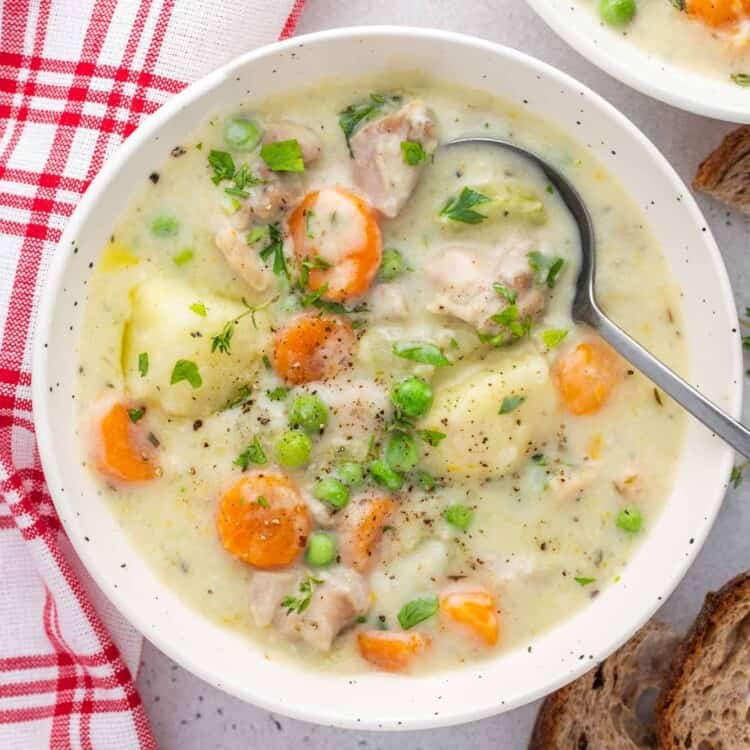 a bowl of creamy chicken stew with peas, potatoes, and carrots, viewed from overhead. there's a spoon in the bowl, and slices of bread next to the bowl.