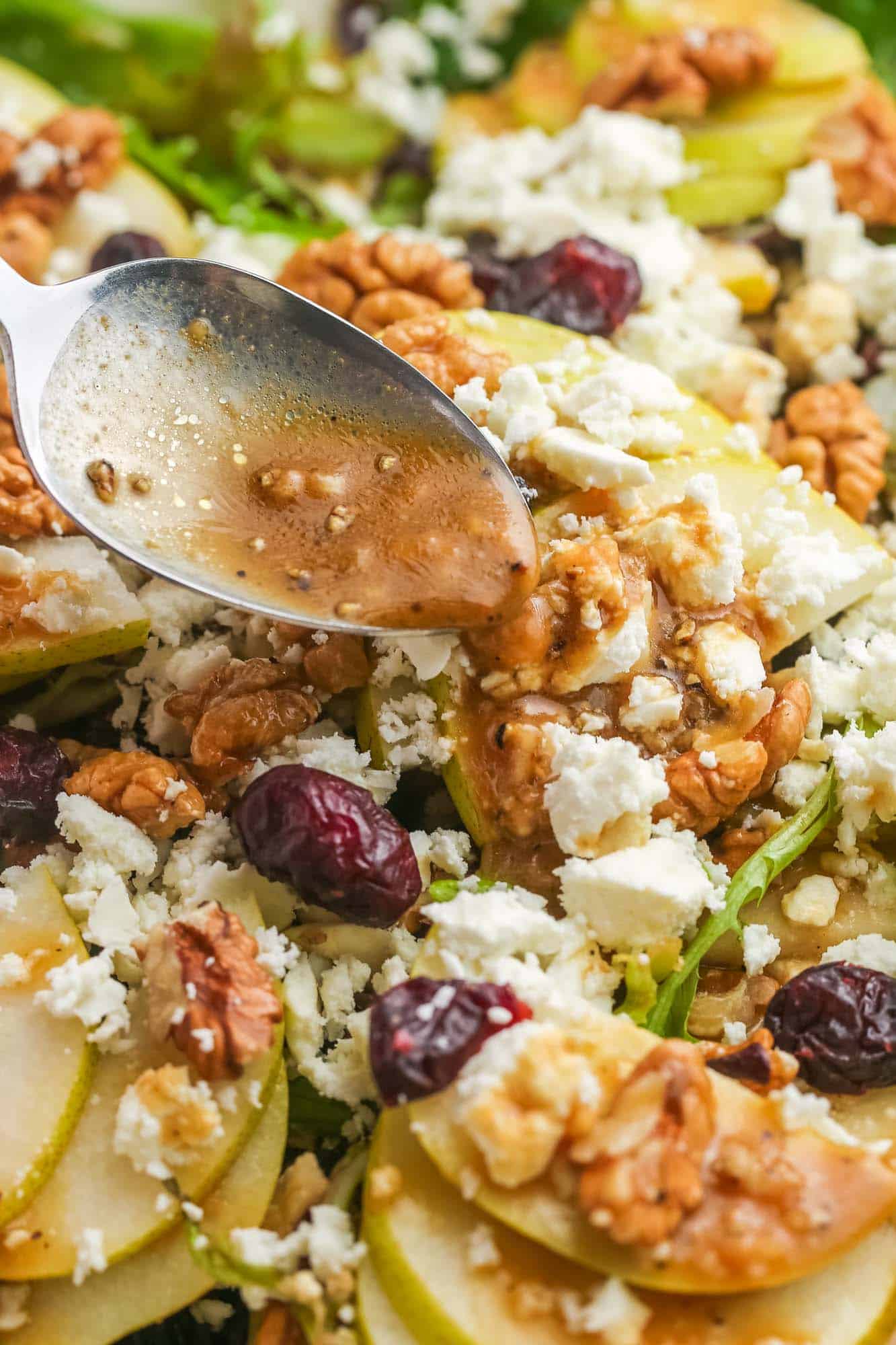 closeup view of a salad topped with sliced pears, cheese, and nuts. A spoon is pouring dressing over the salad.