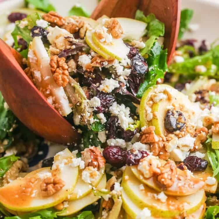 a bowl of salad with pears and nuts, being tossed with two wooden serving spoons