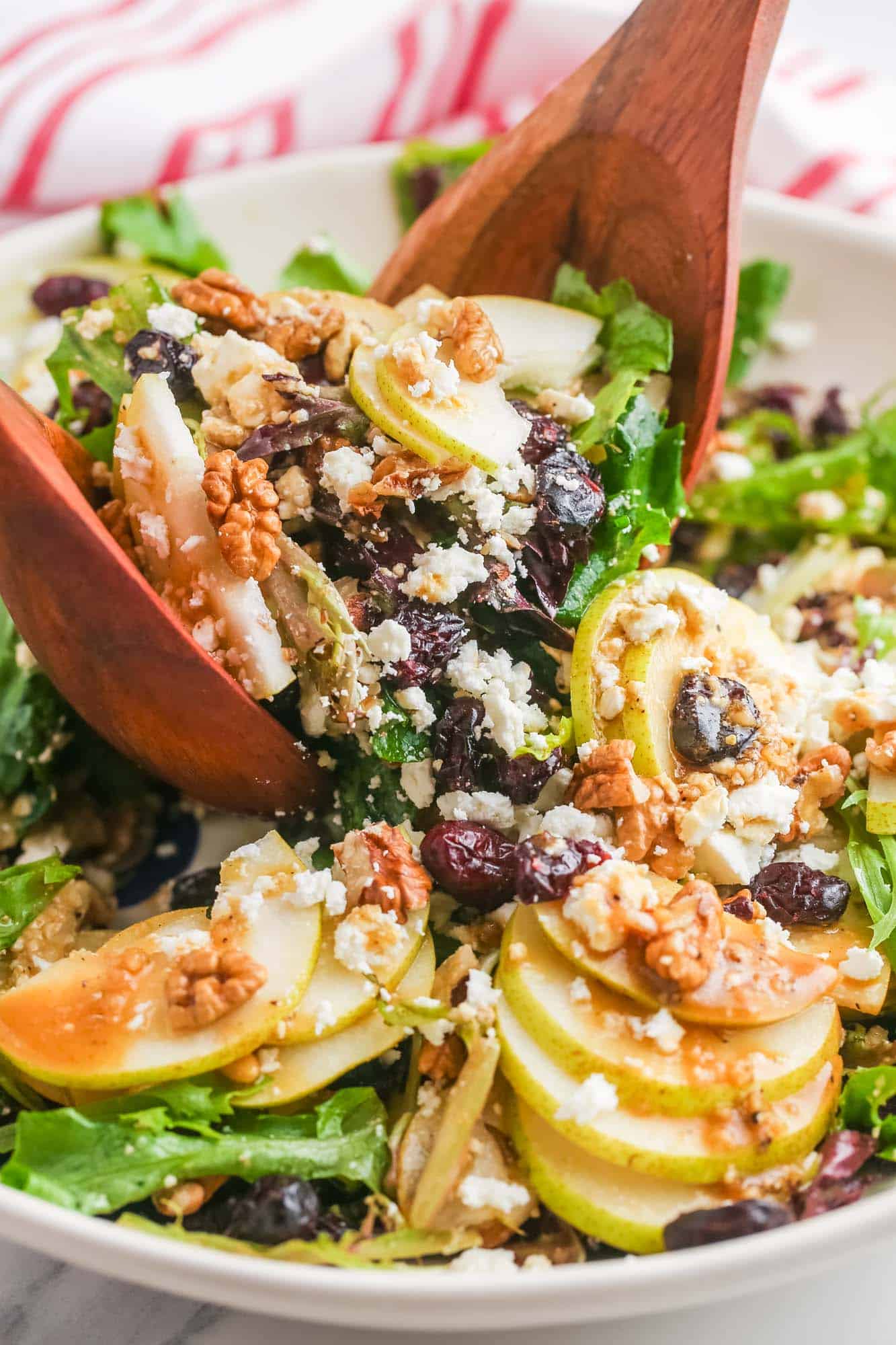 a bowl of salad with pears and nuts, being tossed with two wooden serving spoons