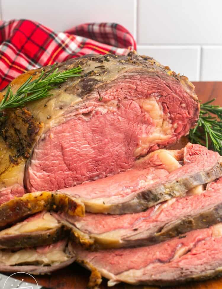A roasted prime rib roast on a wooden cutting board. Slices have been made from the front of it. There is a red plaid towel behind it.