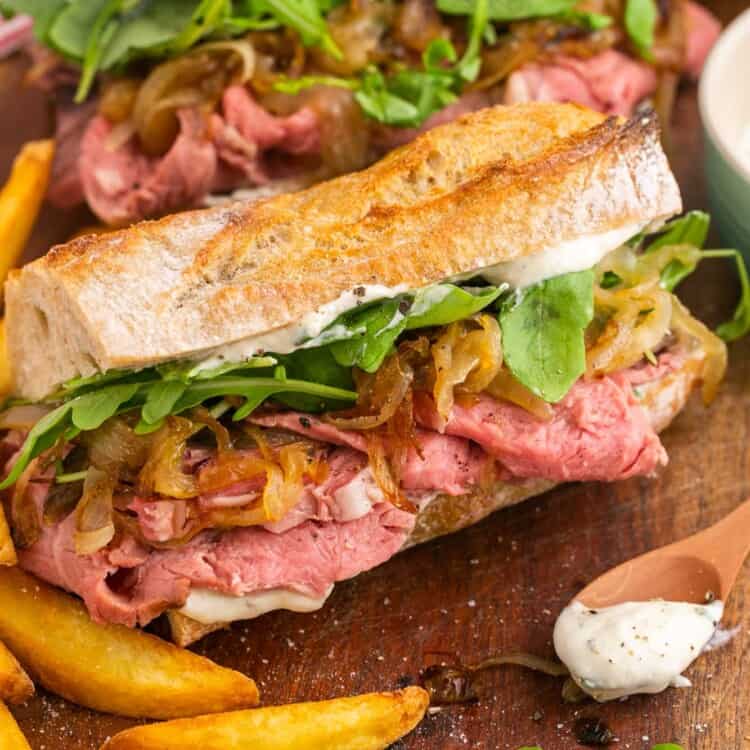 A sandwich made with slided prime rib, onions, and arugula on a baguette. French fries are next to the sandwich. a small wooden spoon with horseradish sauce is on the counter in front of the sandwich on the right.