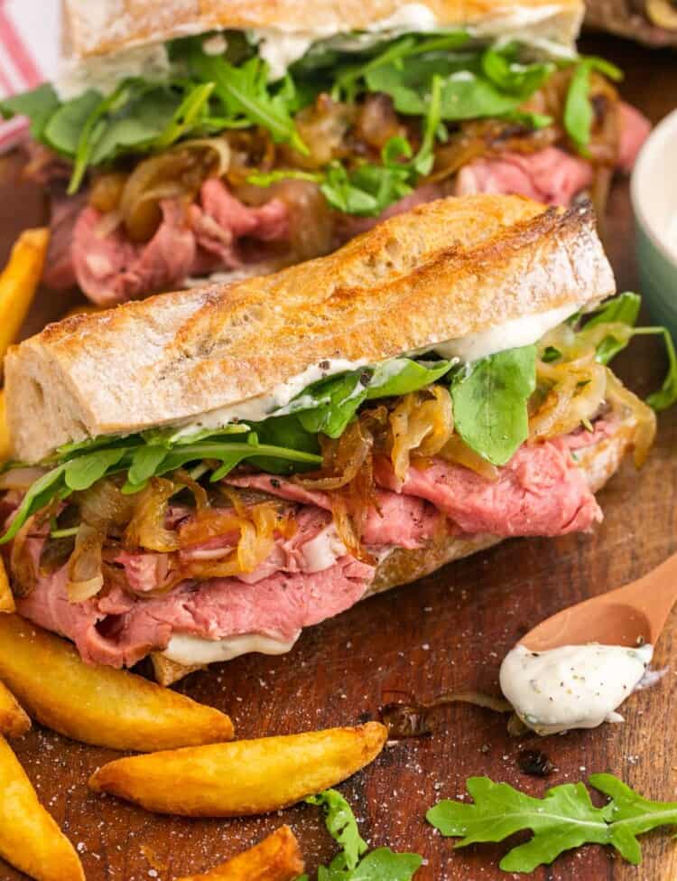 A sandwich made with slided prime rib, onions, and arugula on a baguette. French fries are next to the sandwich. a small wooden spoon with horseradish sauce is on the counter in front of the sandwich on the right.