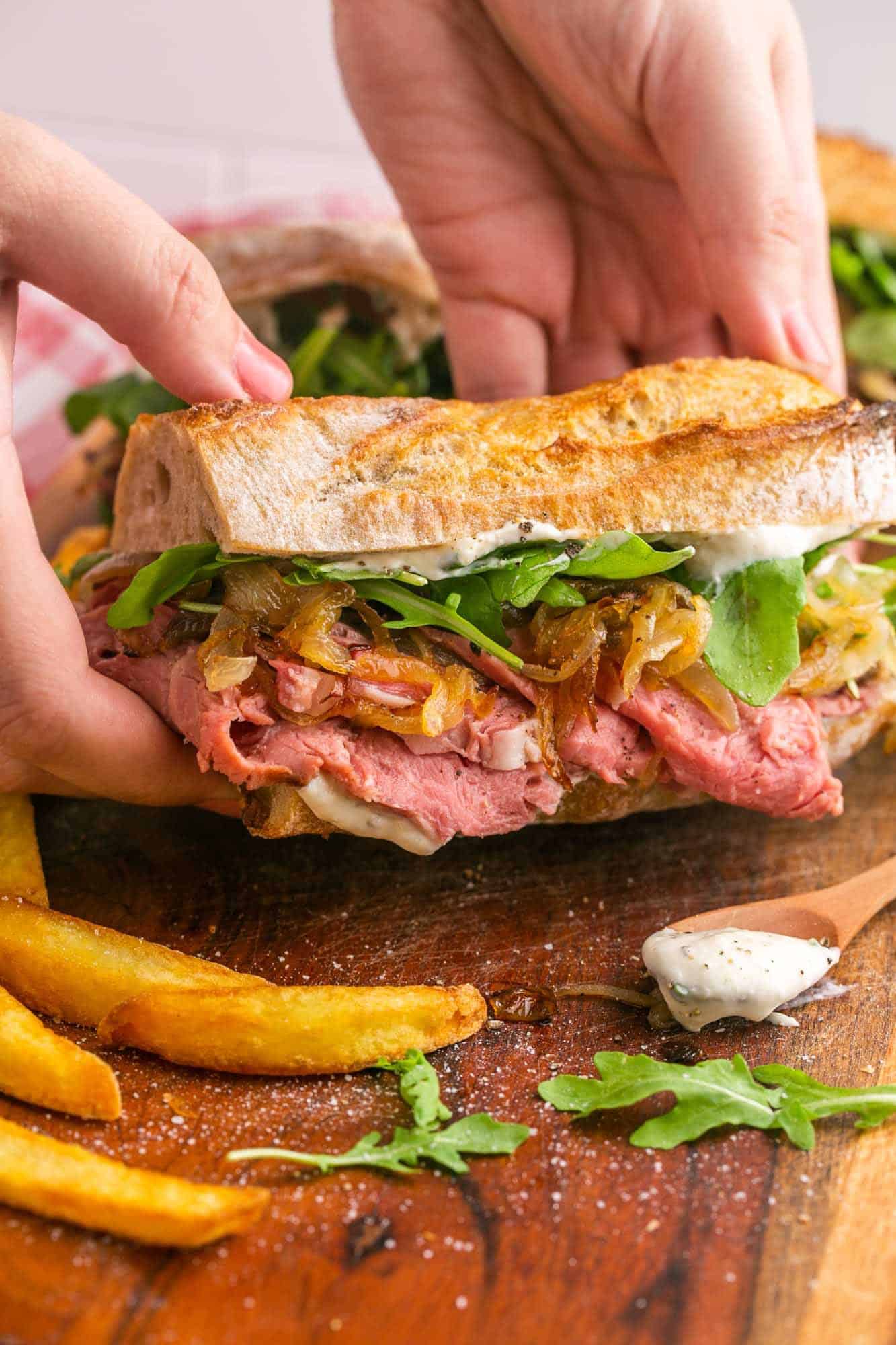 two hands are picking up a prime rib sandwich from a wooden cutting board.