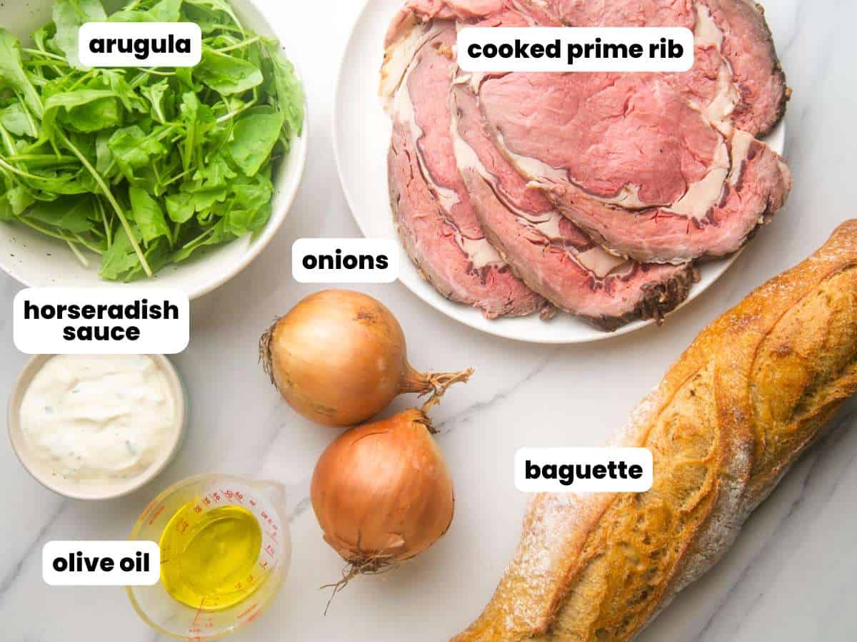 A plate of sliced cooked prime rib, a whole baguette, onions, arugula, horseradish sauce, and olive oil.