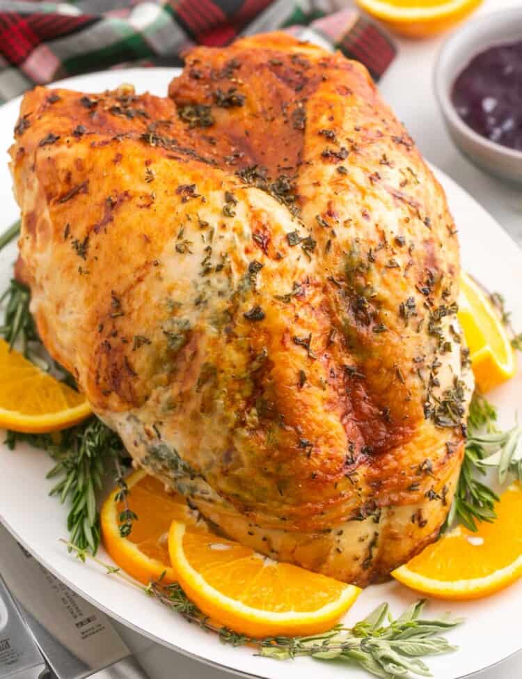 a browned roasted turkey breast on a white platter surrounded by orange slices and herbs.