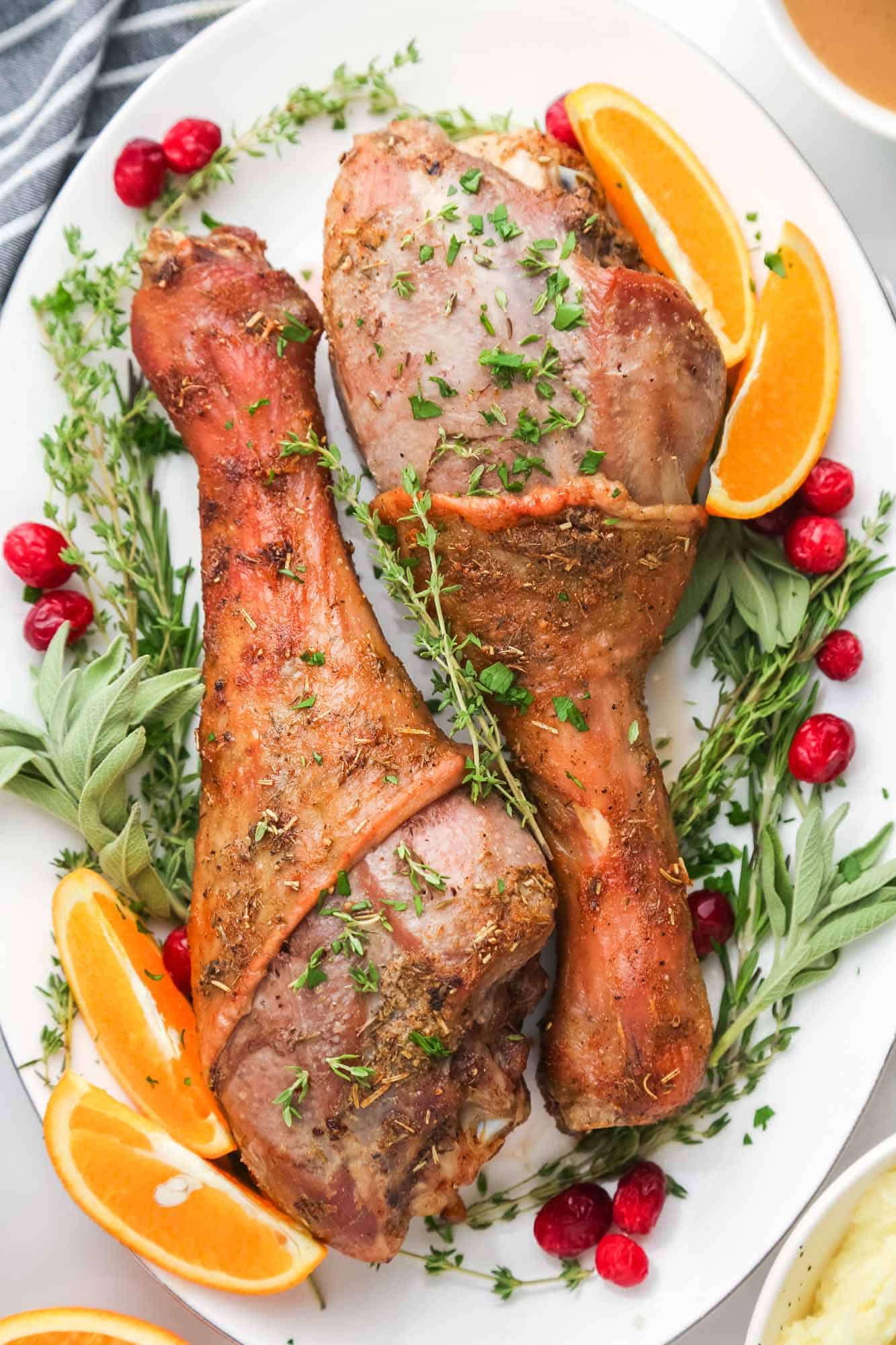Two large roasted turkey legs on a platter, surrounded by cranberries, fresh herbs, and orange wedges.